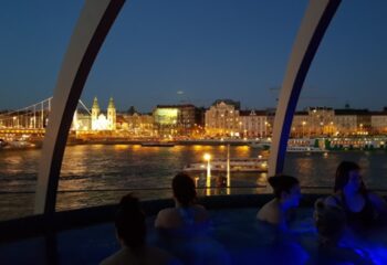 Rudas Thermal Bath Budapest rooftop pool at night. Gay travel advice: Relaxing spa with city view. Danube River, bridge, lights.