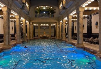 Rudas Thermal Bath, Budapest. Ornate pool hall with columns, a statue, and a man swimming. A gay travel destination for relaxation.