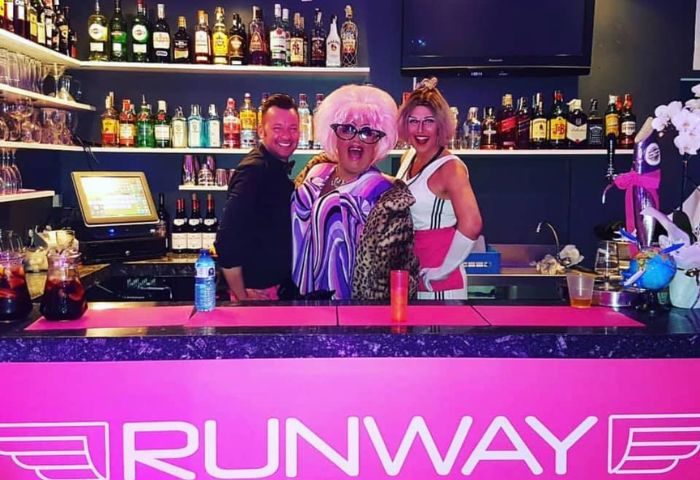 Runway Terrace Bar, a gay bar in Sitges. Staff and drag queens behind the bar with the Runway logo. Gay travel advice.