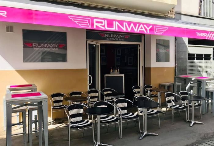 Runway Terrace Bar, a gay bar in Sitges. Exterior view with tables and chairs on the terrace. Pink awning with the bar's name.