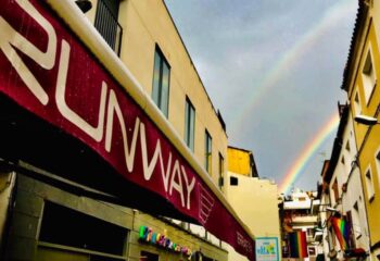 Runway Terrace Bar, a gay bar in Sitges. Rainbows arch over the street, with pride flags visible. A welcoming spot for gay travelers.