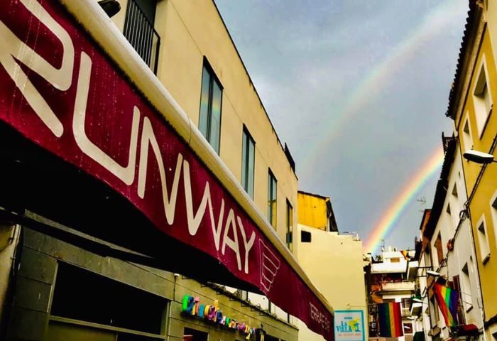 Runway Terrace Bar, a gay bar in Sitges. Rainbows arch over the street, with pride flags visible. A welcoming spot for gay travelers.