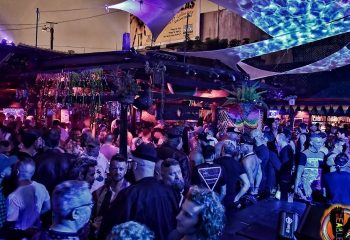 Crowded SF Eagle Bar in San Francisco. Gay bar scene with men socializing under colorful lights & canopies. Popular gay travel destination.