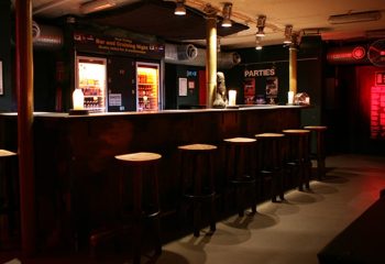 Copenhagen gay bar interior. Dark wood bar with stools, drink fridges, and Parties sign. Gay travel advice in SLM Copenhagen.