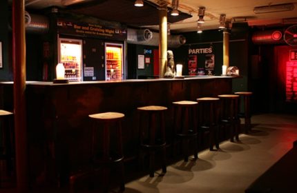 Copenhagen gay bar interior. Dark wood bar with stools, drink fridges, and Parties sign. Gay travel advice in SLM Copenhagen.