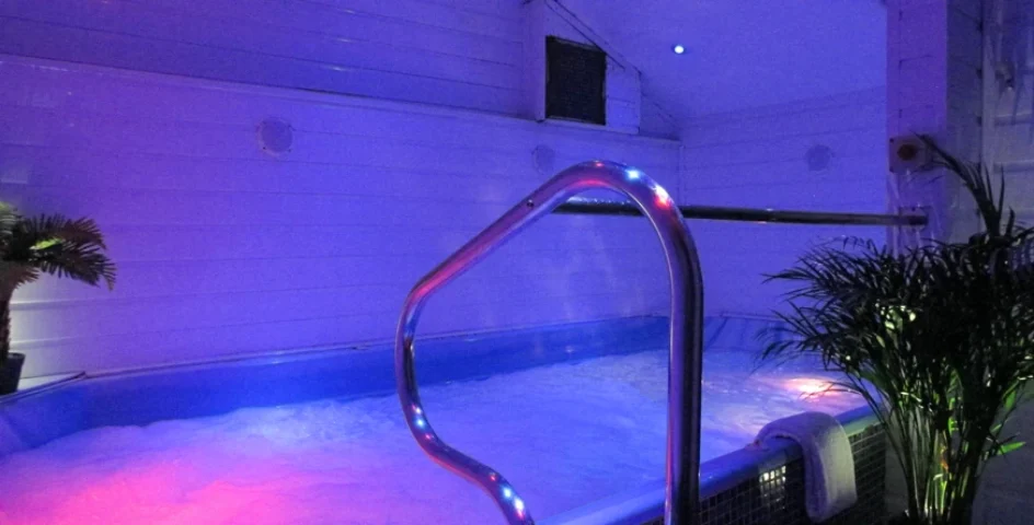 Sailors Sauna gay sauna: Interior view of a hot tub area with blue and pink lighting, a metal handrail, and potted plants, for gay men's travel.