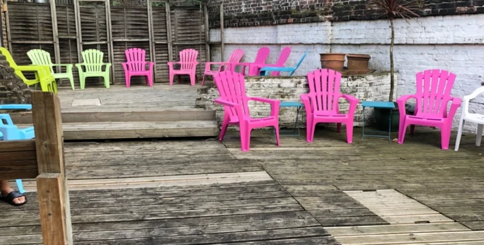 Sailors Sauna: A colorful outdoor area at a gay sauna. Pink, green, and blue chairs are arranged on a wooden deck near a brick wall.