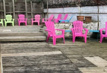 Sailors Sauna: A colorful outdoor area at a gay sauna. Pink, green, and blue chairs are arranged on a wooden deck near a brick wall.
