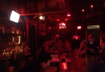 Saloon 1 Key West gay bar in Florida. Red lighting, bar with patrons, and a neon sign that reads C*m Bucket. Nightlife scene.