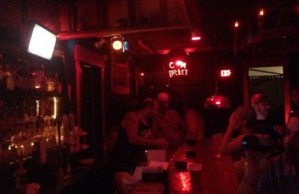 Saloon 1 Key West gay bar in Florida. Red lighting, bar with patrons, and a neon sign that reads C*m Bucket. Nightlife scene.