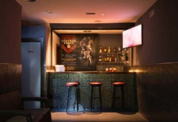 Bar area at Sauna Condal, a gay sauna in Barcelona. Red stools line the bar. A Leather Party poster is displayed.