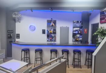 Bar area at a gay sauna in Gran Canaria, Portugal. Blue lights illuminate the bar with stools, shelves of drinks, and a seating area.
