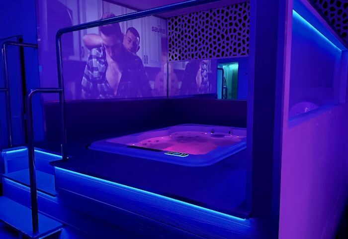 Interior of a gay sauna in Sitges. A hot tub glows with pink light, surrounded by blue LED strip lighting. A screen shows two men embracing.