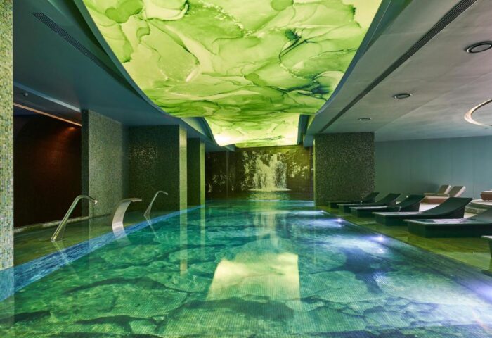 Savoy Palace Madeira pool. Indoor pool with green-toned tile and ceiling art, lounge chairs, and waterfall feature. Gay travel advice.
