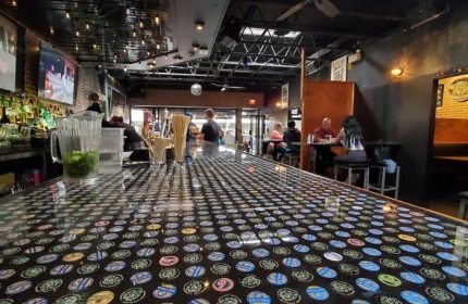Score on Davie bar interior, Vancouver. Bottle cap-covered bar top, patrons at tables. Gay travel advice, sports & nightlife.
