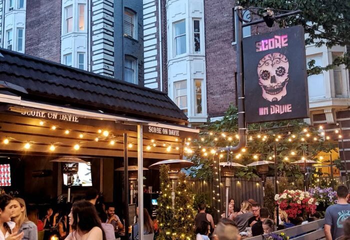 Score on Davie sports bar in Vancouver. Outdoor patio scene with people, lights, heaters, and a sign with a skull. Gay travel advice.