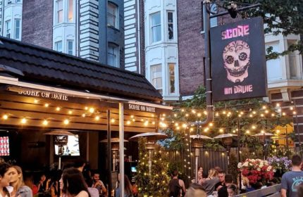 Score on Davie sports bar in Vancouver. Outdoor patio scene with people, lights, heaters, and a sign with a skull. Gay travel advice.
