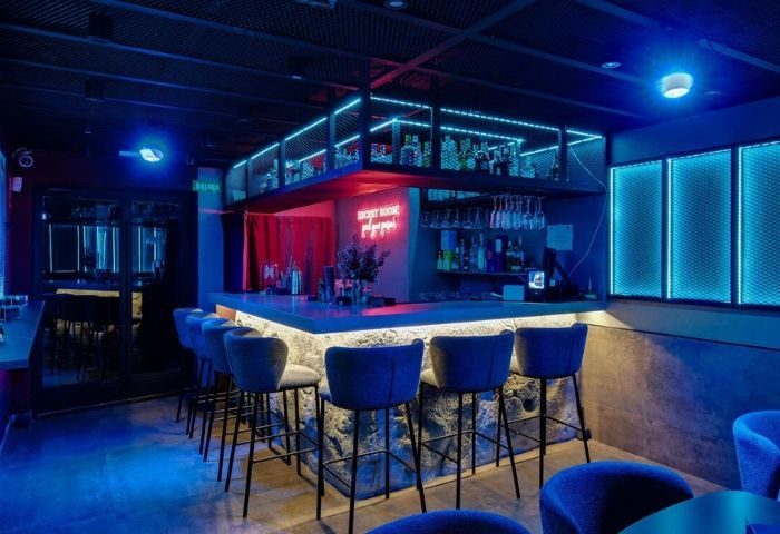Secret Room Bar, Madrid gay bar. Blue neon lights illuminate the bar area with stools, bottles, and a sign. Gay travel.