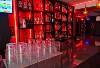 Sensations gay bar in Benidorm. Red lighting illuminates a bar with rows of bottles on shelves. Glasses lined up on the counter. Speakers visible.