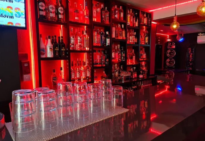 Sensations gay bar in Benidorm. Red lighting illuminates a bar with rows of bottles on shelves. Glasses lined up on the counter. Speakers visible.