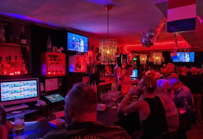 Interior of Sensations gay bar in Benidorm. Crowd at the bar, red lighting, TVs, bottles, and disco ball. Gay travel advice.