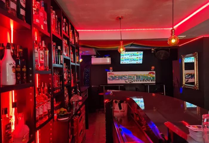 Interior of Sensations, a gay bar in Benidorm. Shelves of liquor bottles glow under red lights, with the bar and logo visible.