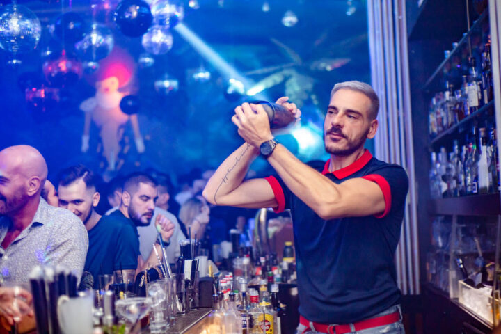 Bartender at Shamone Club, a gay bar, mixes drinks. Disco balls, blue lighting, and a crowd add to the vibrant nightlife atmosphere.