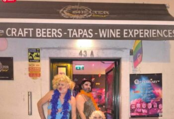 Shelter Bar Lisboa entrance. Drag queens pose outside the gay bar, which offers craft beers, tapas, and wine experiences. Rainbow flag visible.