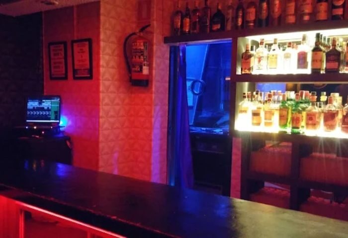 Interior of a gay cruise club bar in Madrid. Shelves of liquor bottles, a shower entrance, and a computer are visible.