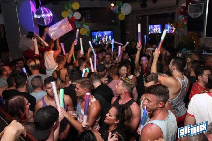Packed Sidetrack gay bar in Chicago. Men hold up glow sticks, disco ball, and TV screens add to the lively atmosphere.