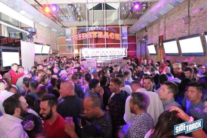 Crowded Sidetrack gay bar in Chicago. Many gay men are standing, drinking, and socializing. Sidetrack sign visible. Gay travel advice.