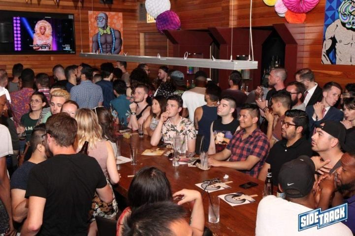 Sidetrack Gay Bar in Chicago is crowded. People are standing and sitting at the bar with drinks. TV and art on the walls.