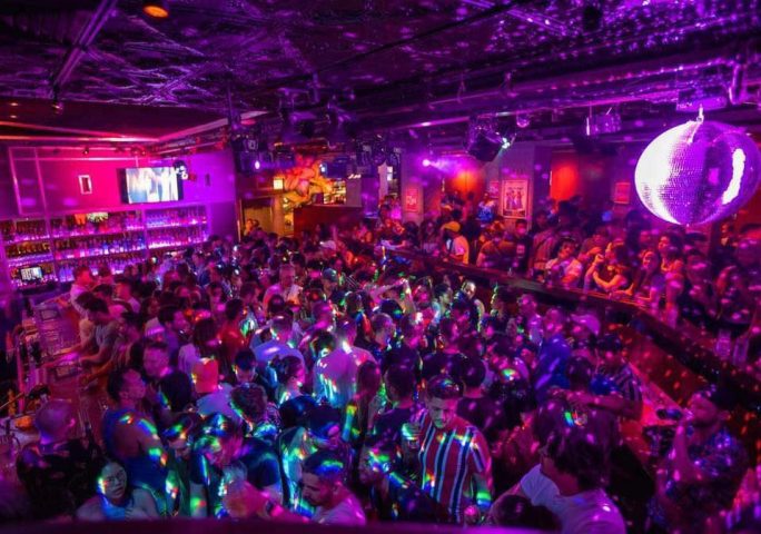 Sidetrack gay bar interior. A packed crowd of men under purple lights and disco ball. Gay travel nightlife scene. Cheerful, fun atmosphere.