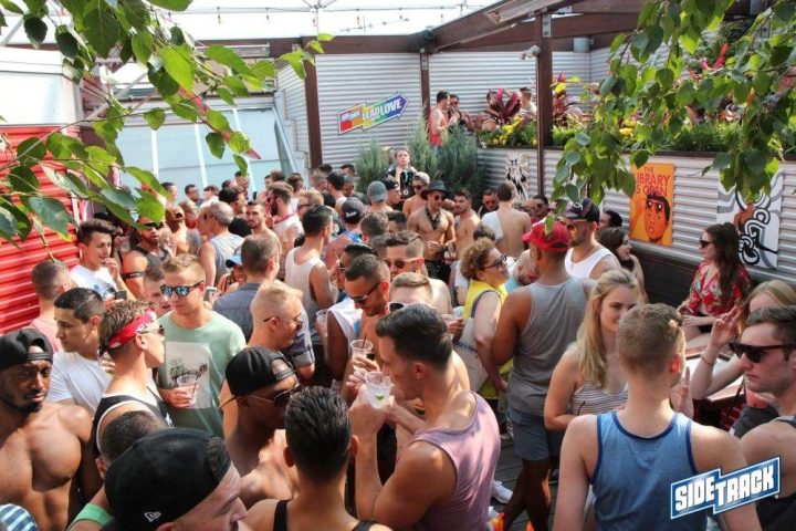 Crowded outdoor patio at Sidetrack gay bar. Men and women socialize, some shirtless, under a leafy trellis. Lead Love sign visible.
