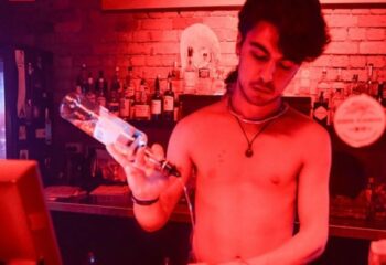 Shirtless bartender pouring a drink at Sircuit Bar, Melbourne. Gay travel nightlife scene with red lighting and liquor bottles in the background.