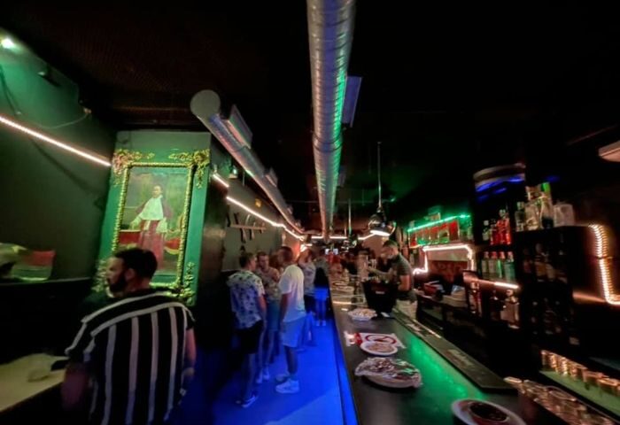 Sixta Bar, a gay bar in Madrid. People line up at the bar in a dimly lit space with green and blue neon lights. Portrait on the wall.