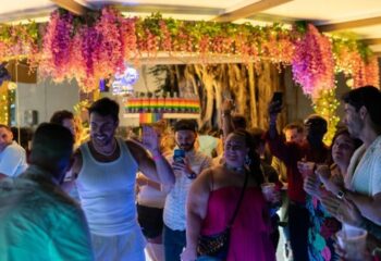 Sky Bar at Axel Hotel, Barcelona. Gay bar scene with people dancing and socializing under floral decorations and rainbow bottles.