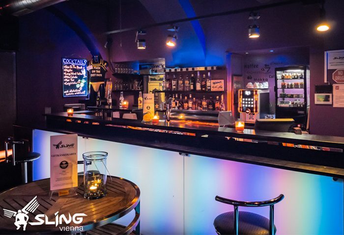 Interior of Sling Vienna, a gay bar. Bar with colorful lights, bottles, and cocktail menu. Table with candle and Sling logo.