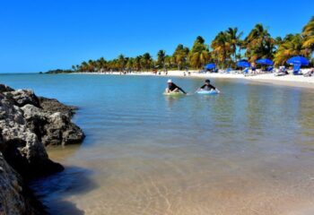 Gay cruise day at Smathers Beach in Key West. People enjoy the clear water, palm trees, and white sand. Blue umbrellas dot the shoreline.