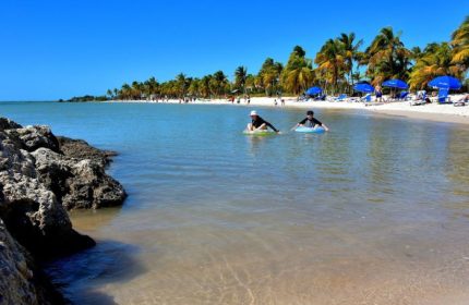 Gay cruise day at Smathers Beach in Key West. People enjoy the clear water, palm trees, and white sand. Blue umbrellas dot the shoreline.