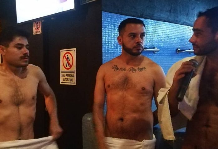 Gay men at Sodome Bath House. Three shirtless men, one with a microphone, stand in a bathhouse setting. Gay travel and bathhouse culture.