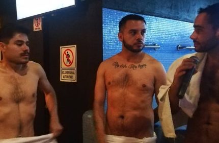 Gay men at Sodome Bath House. Three shirtless men, one with a microphone, stand in a bathhouse setting. Gay travel and bathhouse culture.