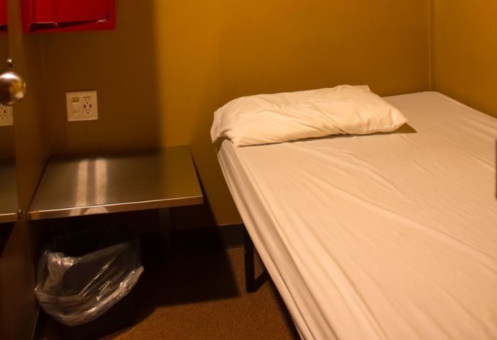 Toronto spa excess: Simple bed with white linens and pillow, bedside table with trash can. Budget-friendly accommodation for gay travelers.