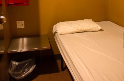 Toronto spa excess: Simple bed with white linens and pillow, bedside table with trash can. Budget-friendly accommodation for gay travelers.