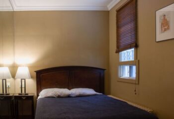 Toronto spa excess: Bedroom with a double bed, bedside tables with lamps, window with blinds, and a framed gay-themed picture on the wall.