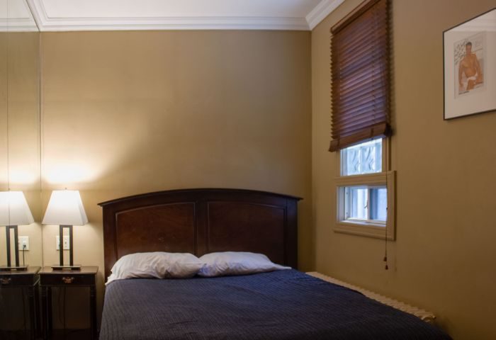Toronto spa excess: Bedroom with a double bed, bedside tables with lamps, window with blinds, and a framed gay-themed picture on the wall.