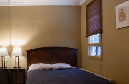 Toronto spa excess: Bedroom with a double bed, bedside tables with lamps, window with blinds, and a framed gay-themed picture on the wall.