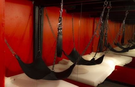 Toronto's Spa Excess: A bed with black sling, chains, and leather restraints. Red walls and mirrored background. Gay travel advice.