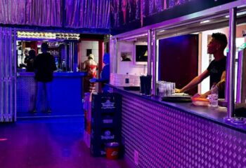 Inside Sparkles Show Bar, a gay bar in Gran Canaria. Bartender serves drinks under purple lights. A vibrant nightlife scene.