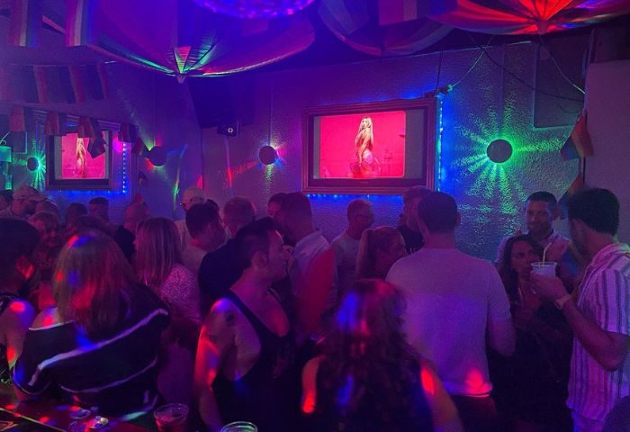 Spirit Bar gay party in Benidorm, Spain. Crowded gay bar with people socializing under colorful lights, rainbow flags, and a TV screen.